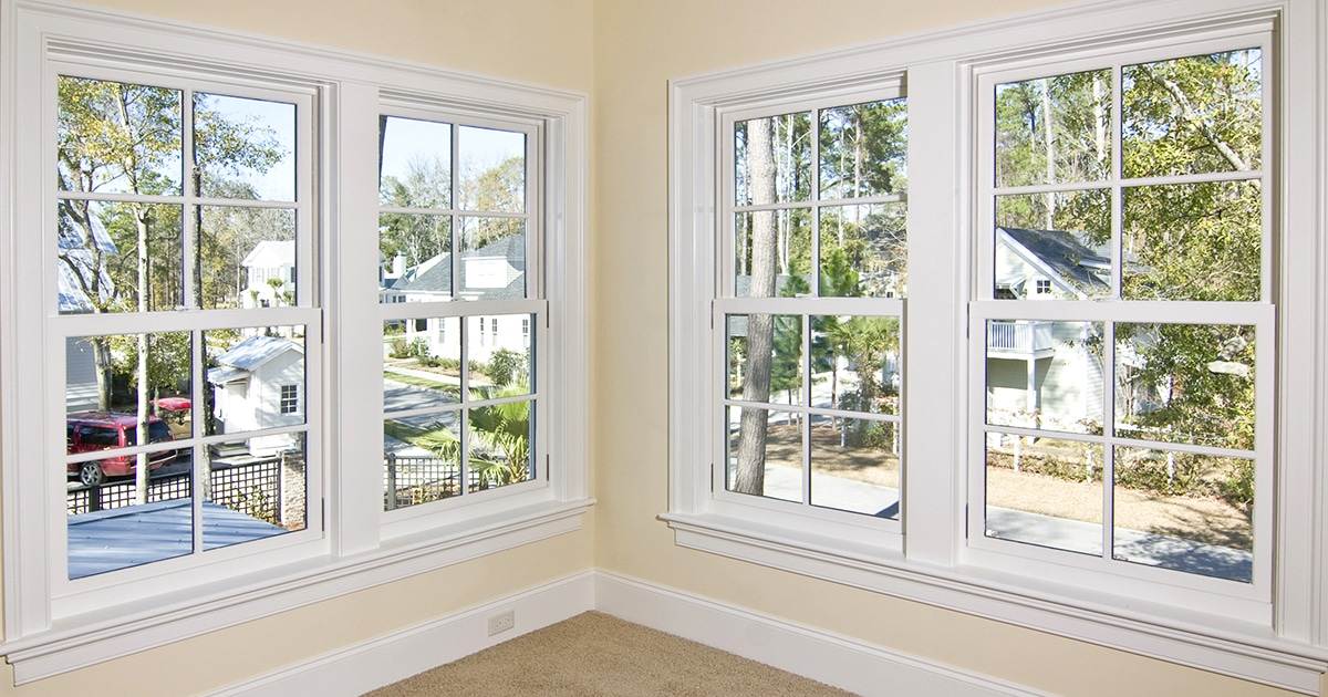 Corner of a room with two sets of white-framed double-hung windows, perfect for those considering buying windows. These overlook neighboring houses and trees. Walls are light-colored and the floor is carpeted.
