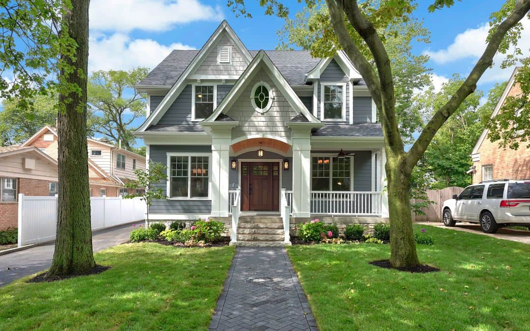 A charming two-story house with a gray exterior and white trim, featuring a front porch and a stone pathway, nestled between lush green trees.