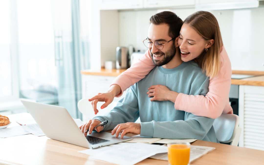 A young couple happily using a laptop together in a bright kitchen, with drinks and a pastry on the table.