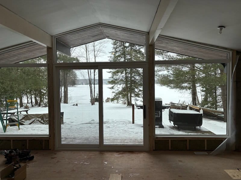 AFTER 1 Large glass windows in a room overlook a snowy landscape with trees, a lake, and outdoor equipment, including a grill and a ladder.