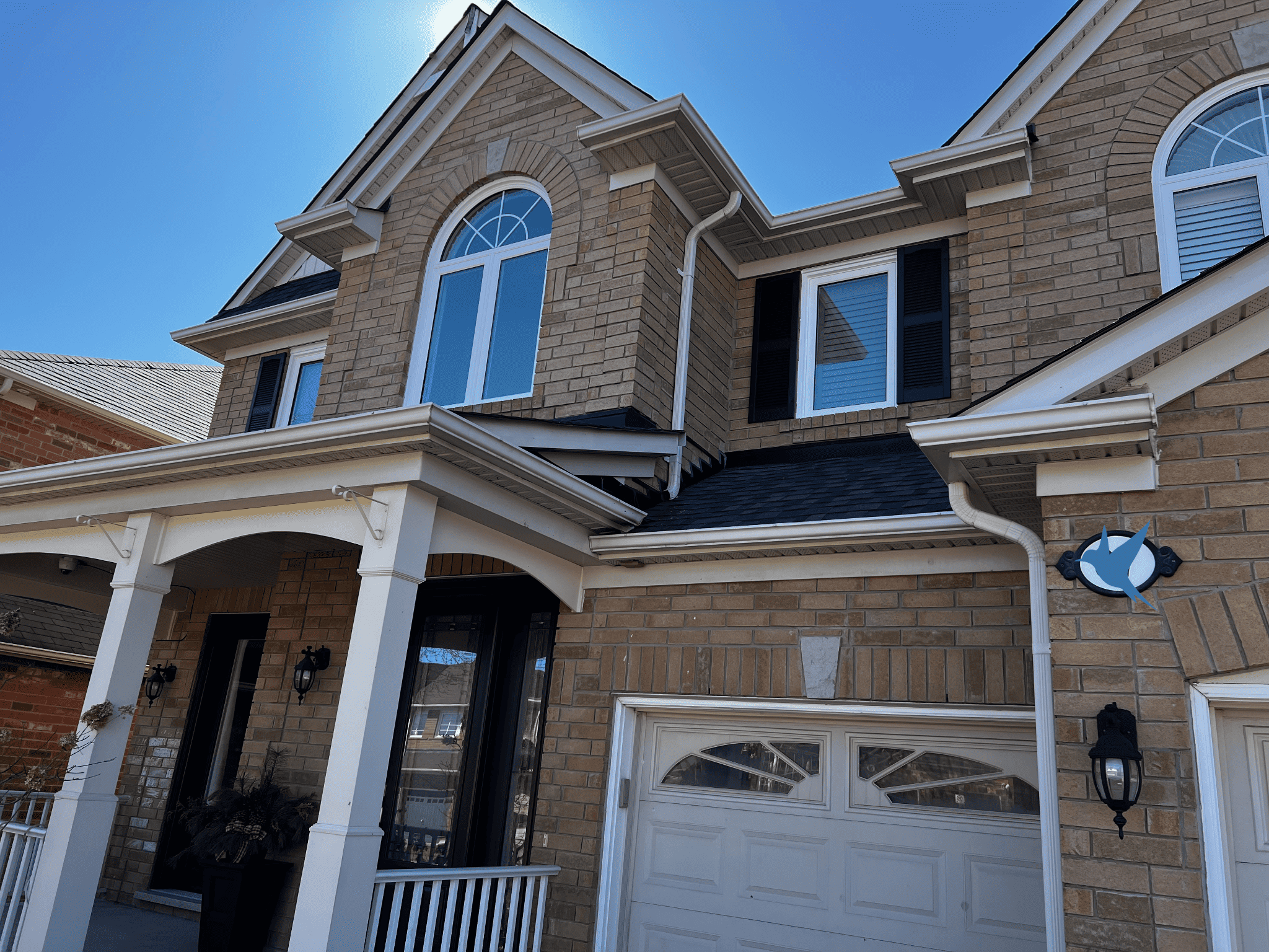A Two-Story Brick House With Arched Windows, A Covered Porch, And A White Garage Door Under Clear Blue Skies.