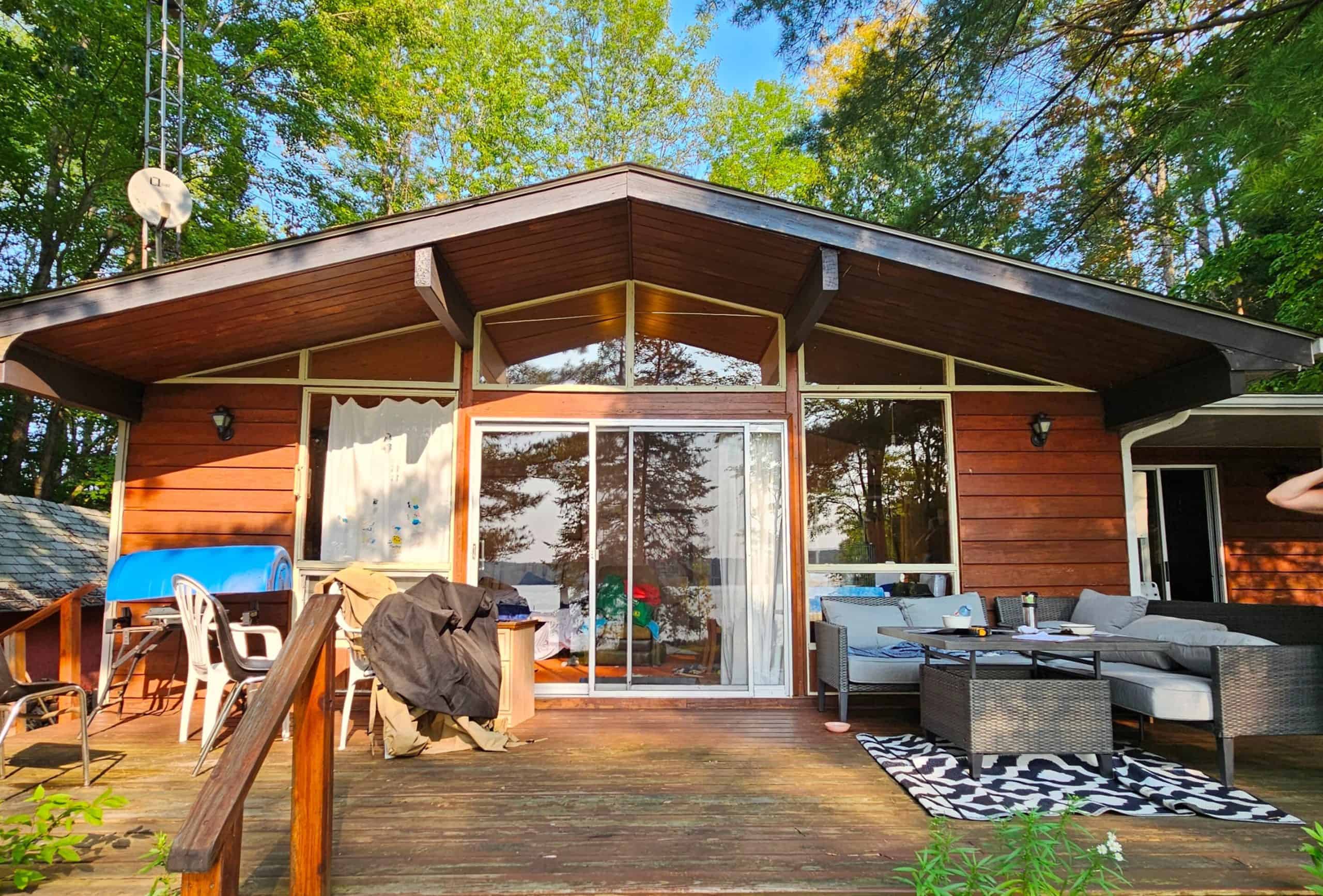 before 2 A rustic cabin with a wooden deck, sliding glass doors, patio furniture, and a grill, surrounded by trees under a clear blue sky.