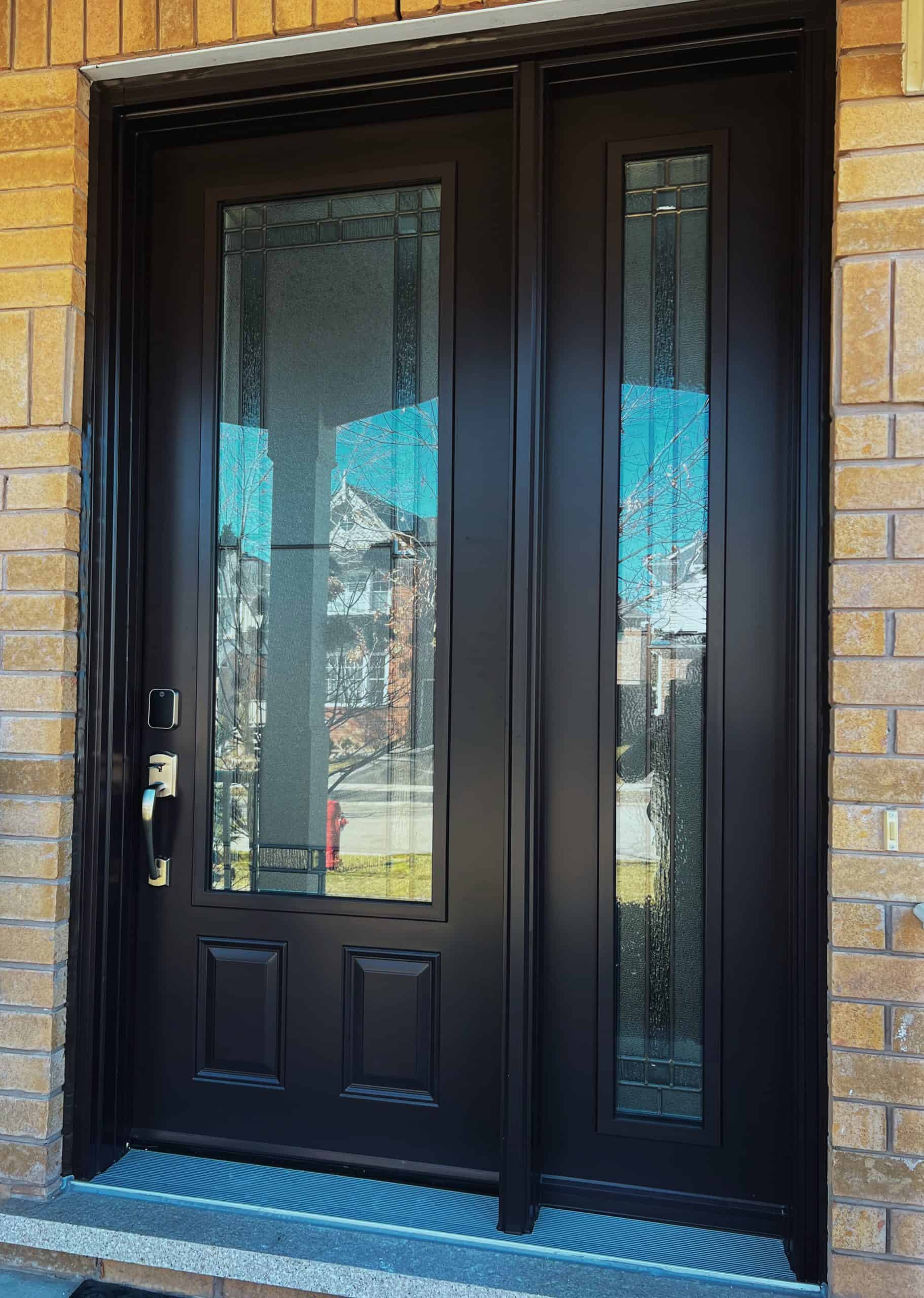 Black Front Door With Two Vertical Glass Panels And A Silver Handle, Set Against A Light Brown Brick Exterior. Reflections Of Houses And Trees Are Visible In The Glass.