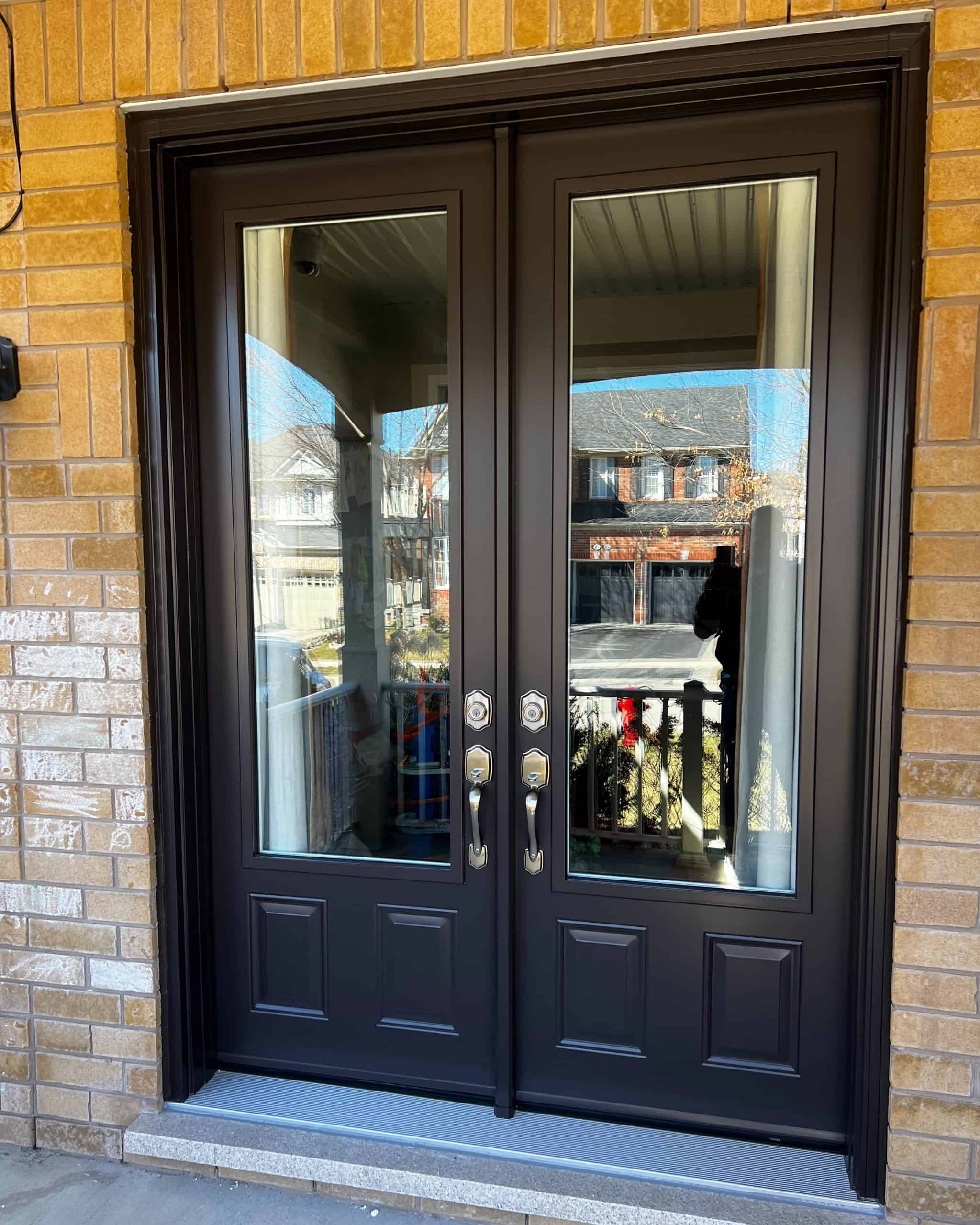 A Set Of Double Black Doors With Large Glass Panels, Surrounded By A Brick Wall, Reflecting Nearby Houses.