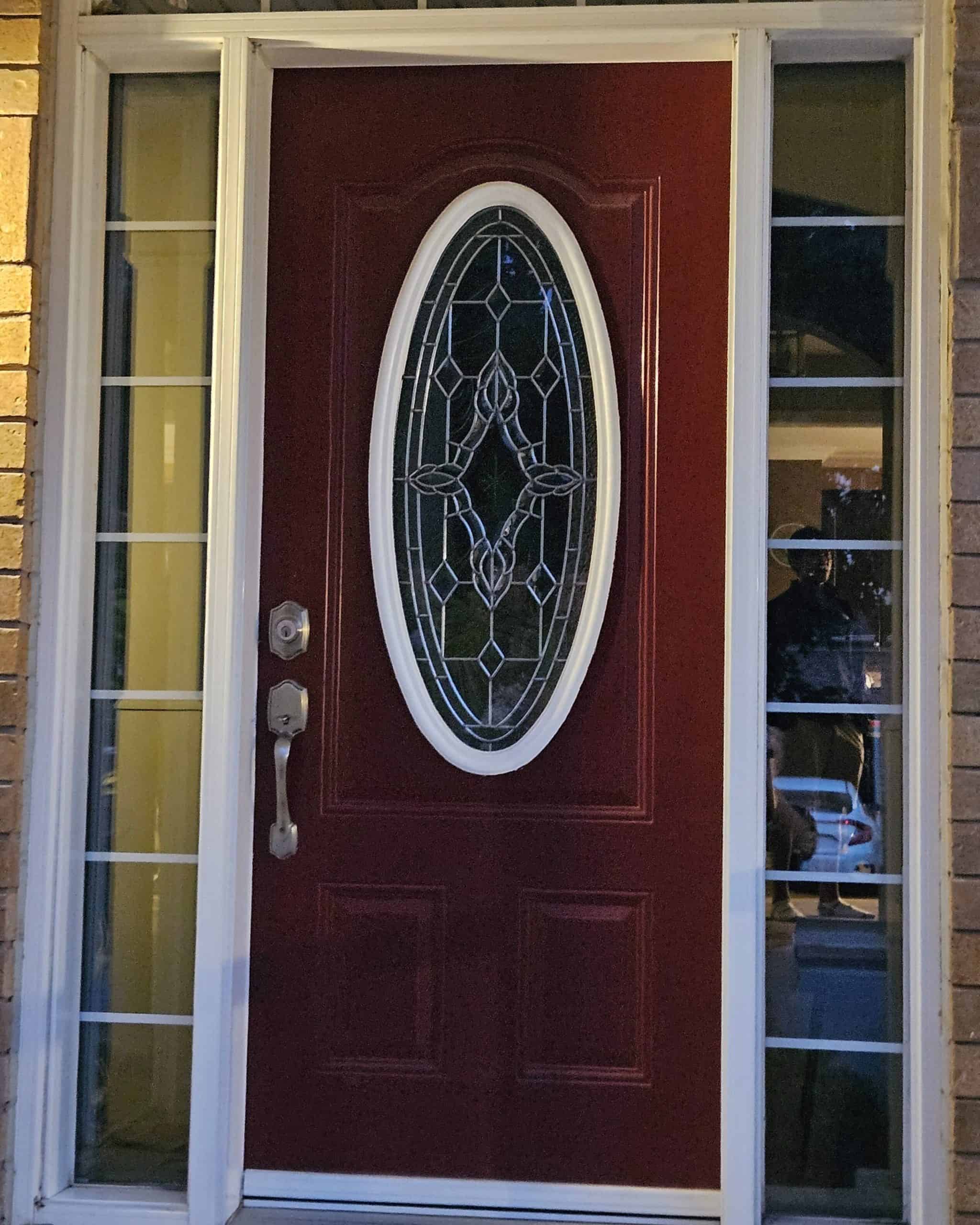 Red Front Door With An Oval Glass Design In The Center And Flanked By Narrow Side Windows, Set In A Brick Facade.