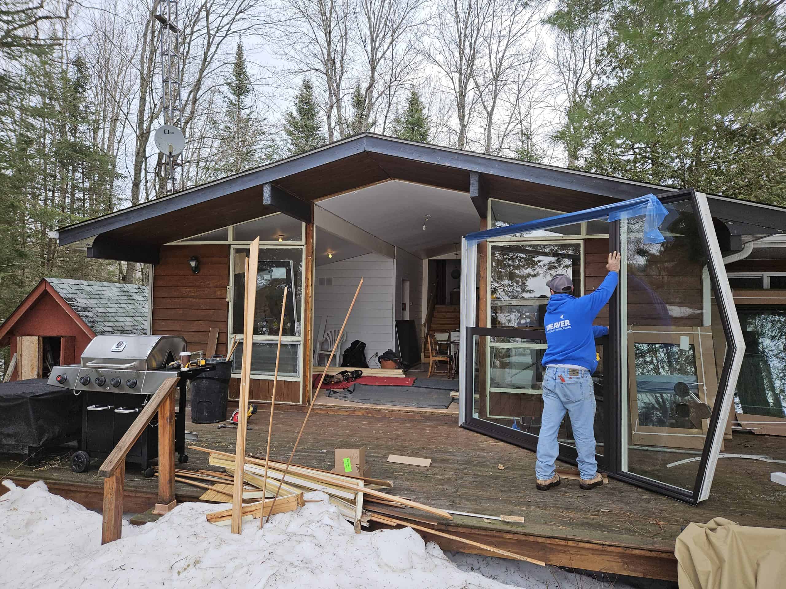 in progress 2 A worker installs a large window on a wooden cabin. The deck is cluttered with construction materials, and a grill is visible. Snow covers the ground, and trees surround the cabin.