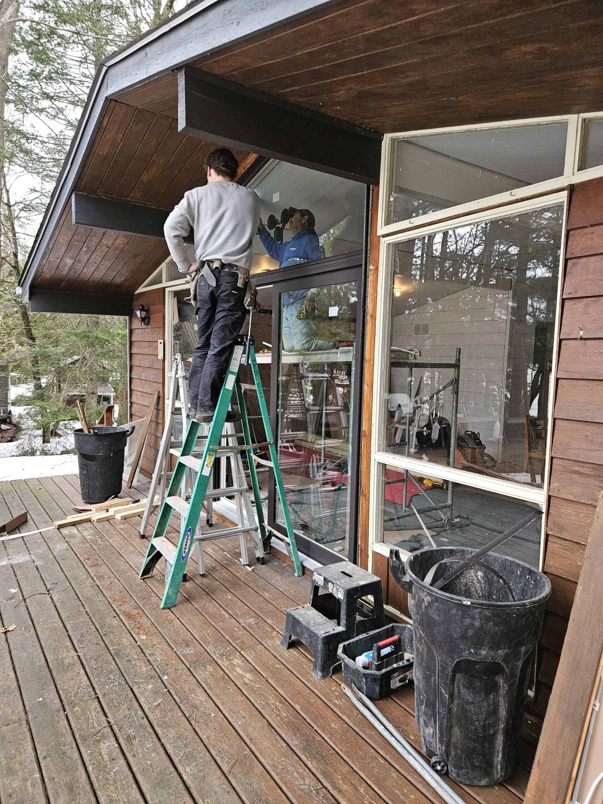 in progress Two people are working on a house exterior; one is on a green ladder, the other is by a window. Tools and a black trash can are on the wooden deck.