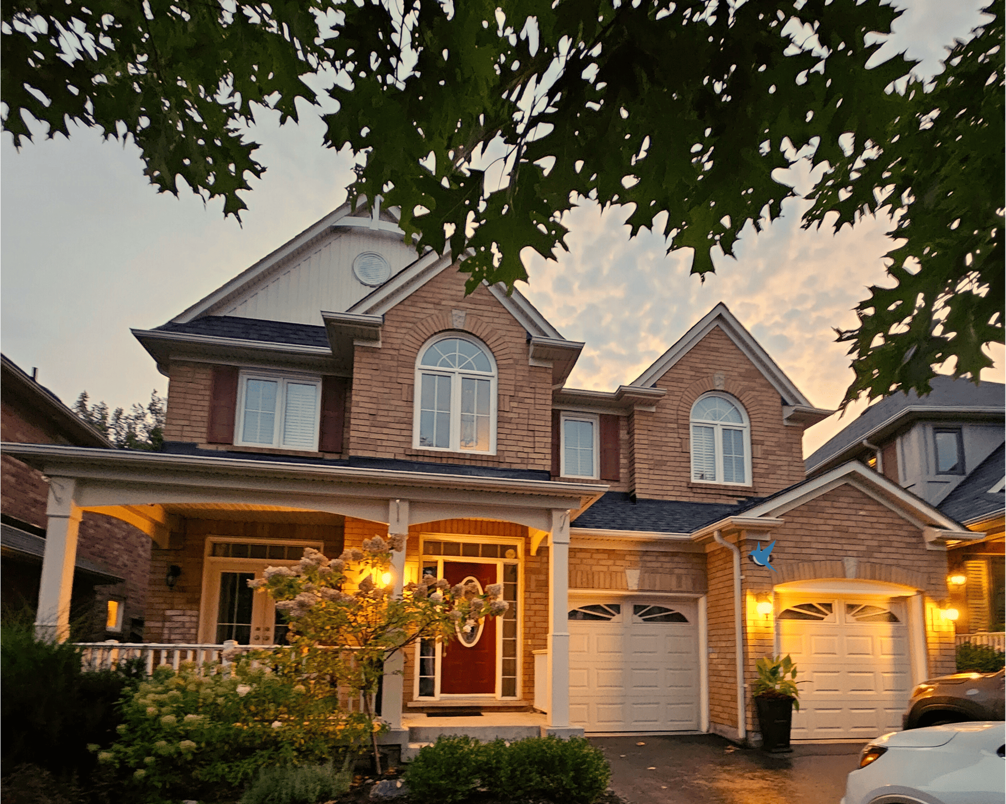Two-Story Brick House With A Front Porch And Two White Garage Doors. A Tree Frames The View, And The Sky Shows A Mix Of Light Clouds During Sunset. A Car Is Partially Visible On The Right.