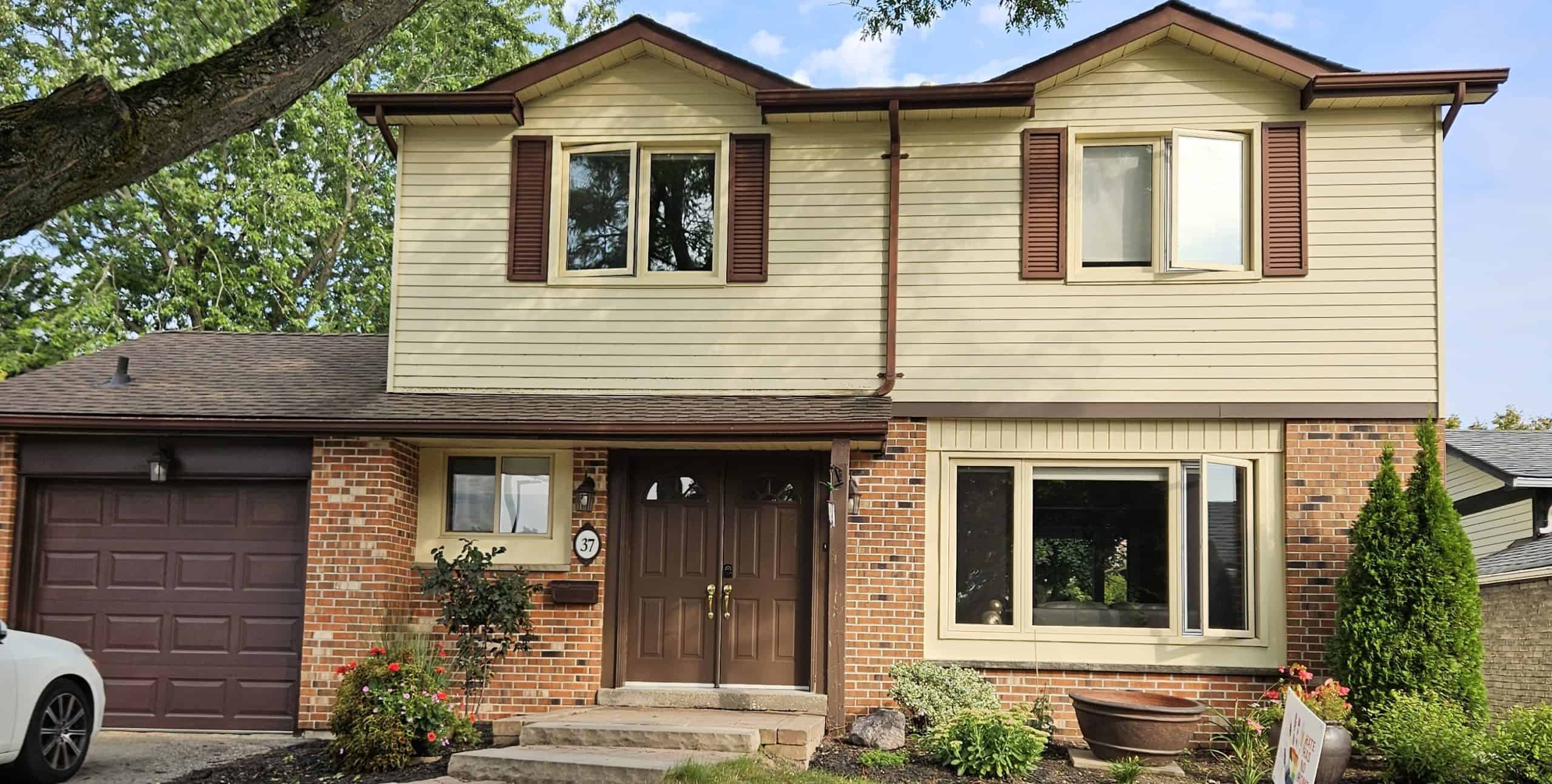 Two-story suburban house with brick and beige siding, brown shutters, double front doors, and attached garage; landscaped front yard with bushes and flowers.