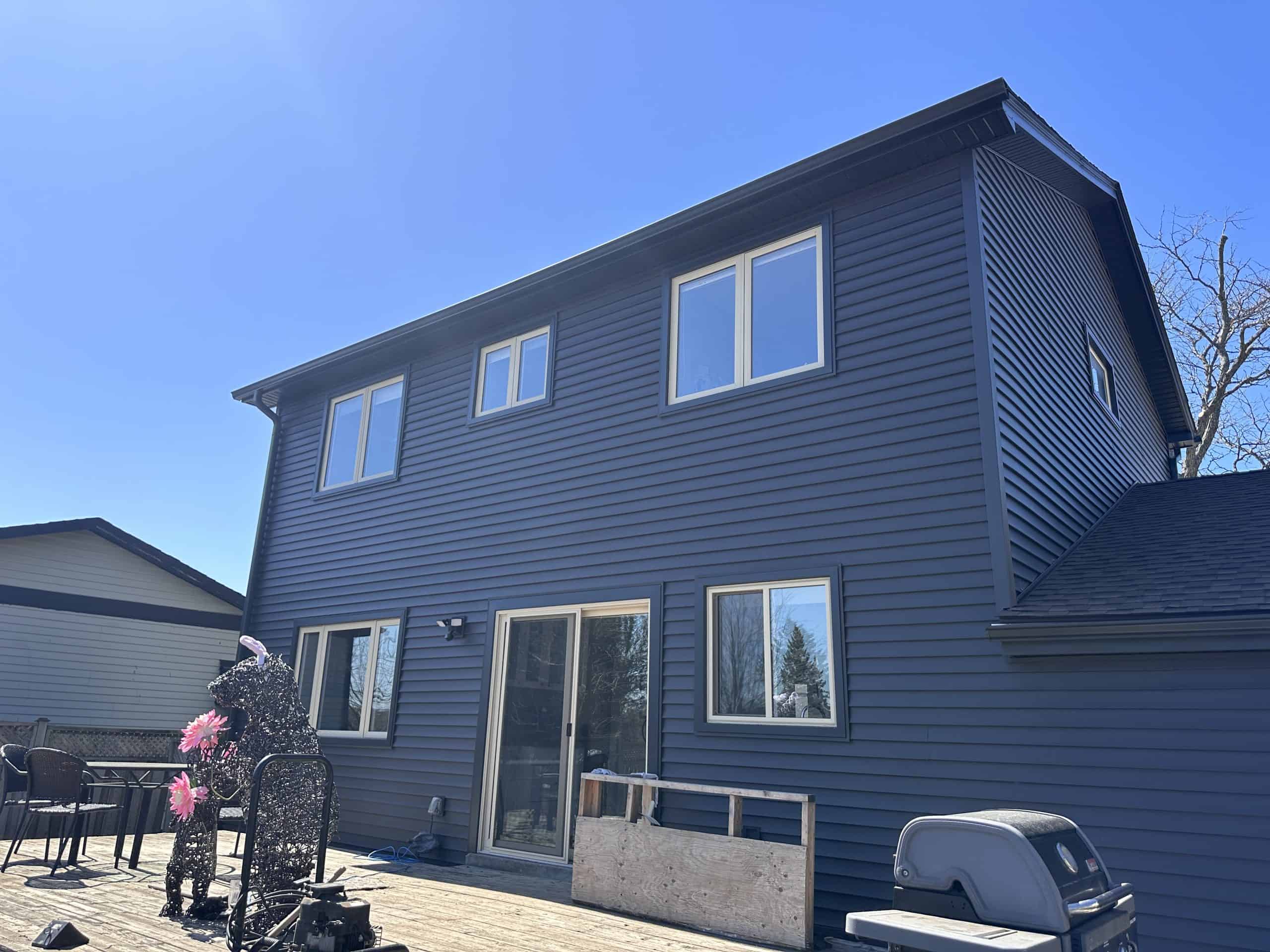 A two-story house with dark blue siding, several windows, a sliding glass door, a grill, patio furniture, and a metal sculpture on a wooden deck under a clear blue sky.