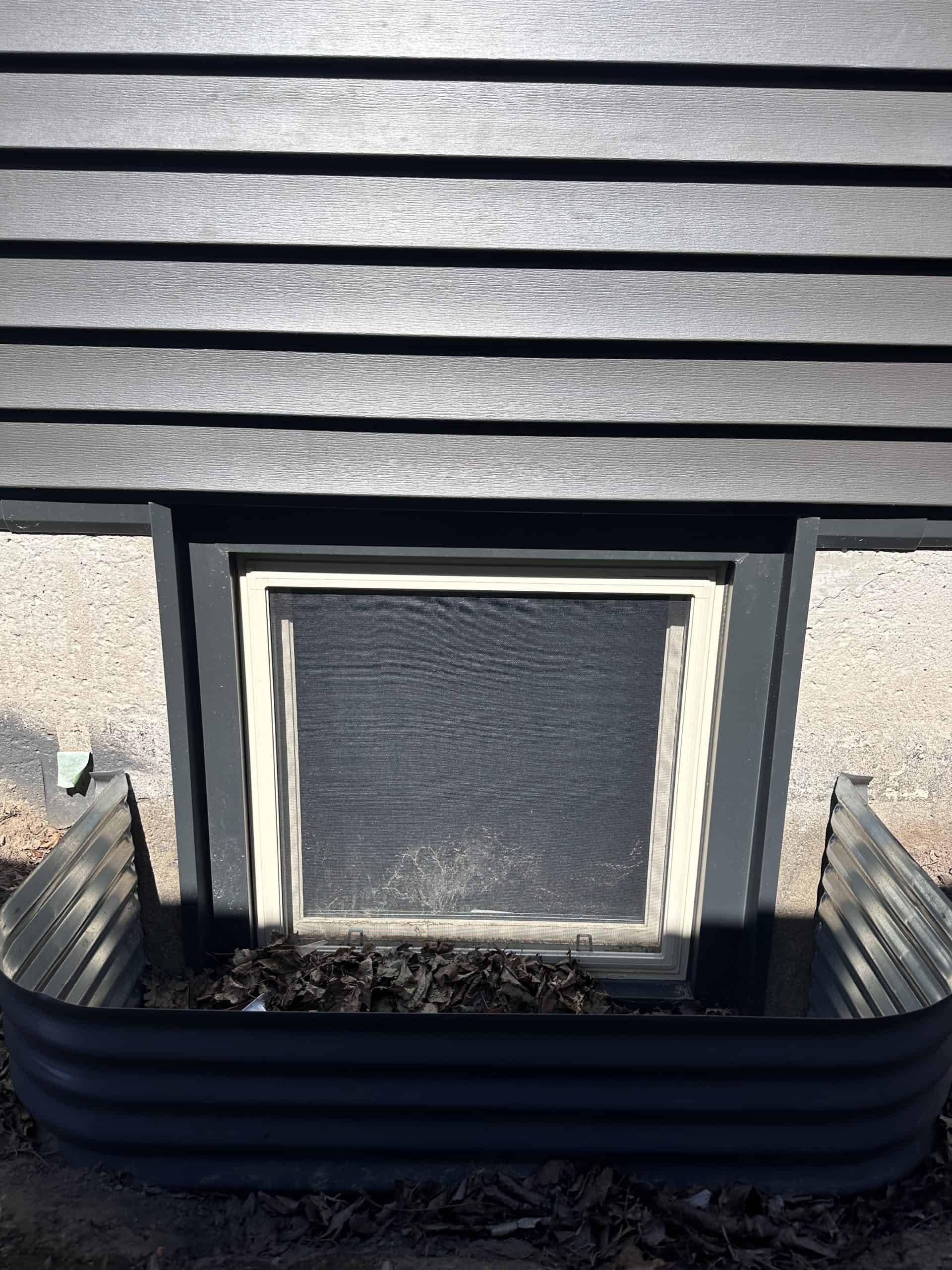 A basement window with a metal window well, surrounded by dark horizontal siding and a concrete foundation, with leaves collected at the bottom.