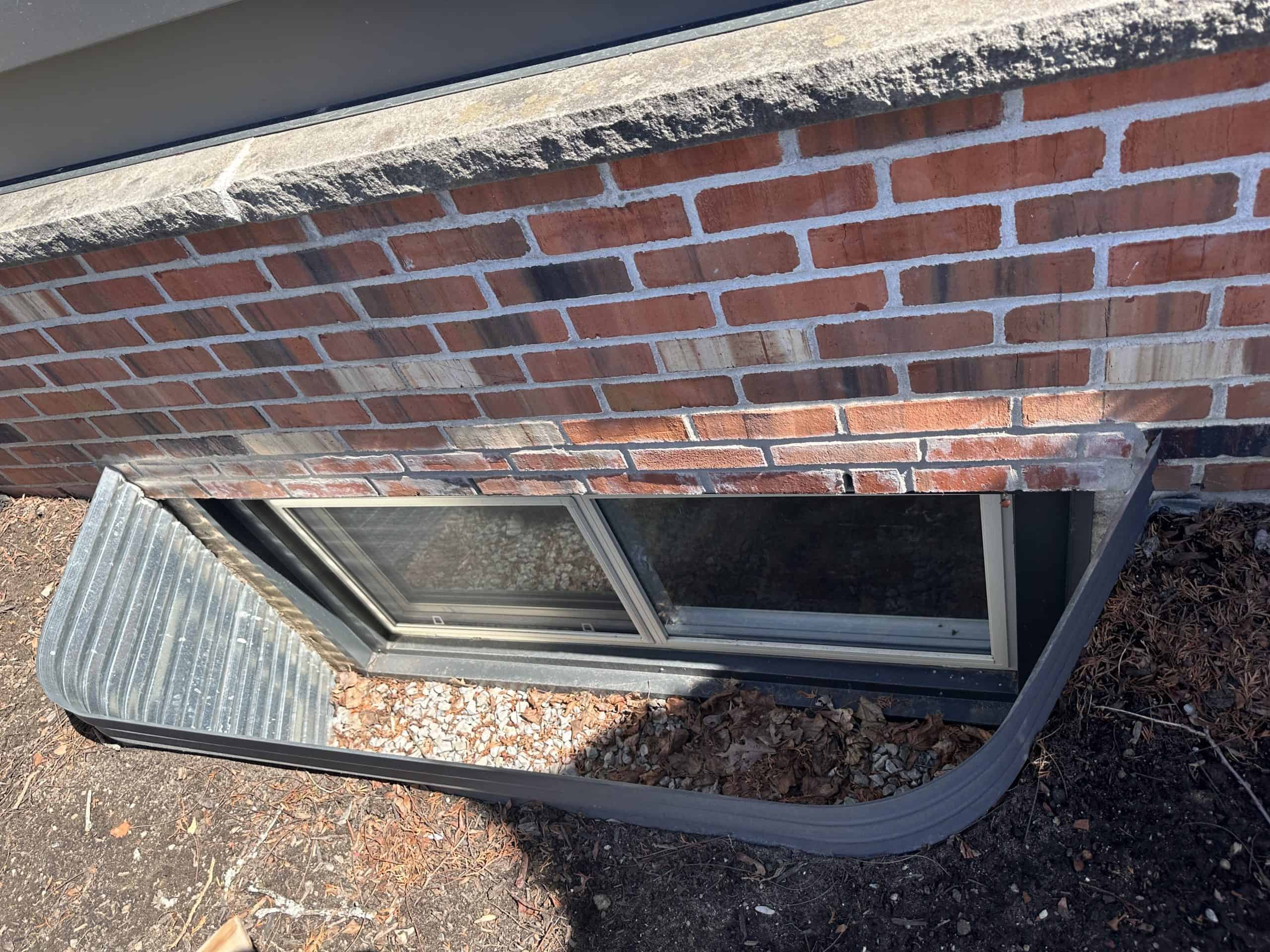 A ground-level basement window with a metal window well, scattered leaves and debris inside, set in a brick wall with a concrete ledge above.