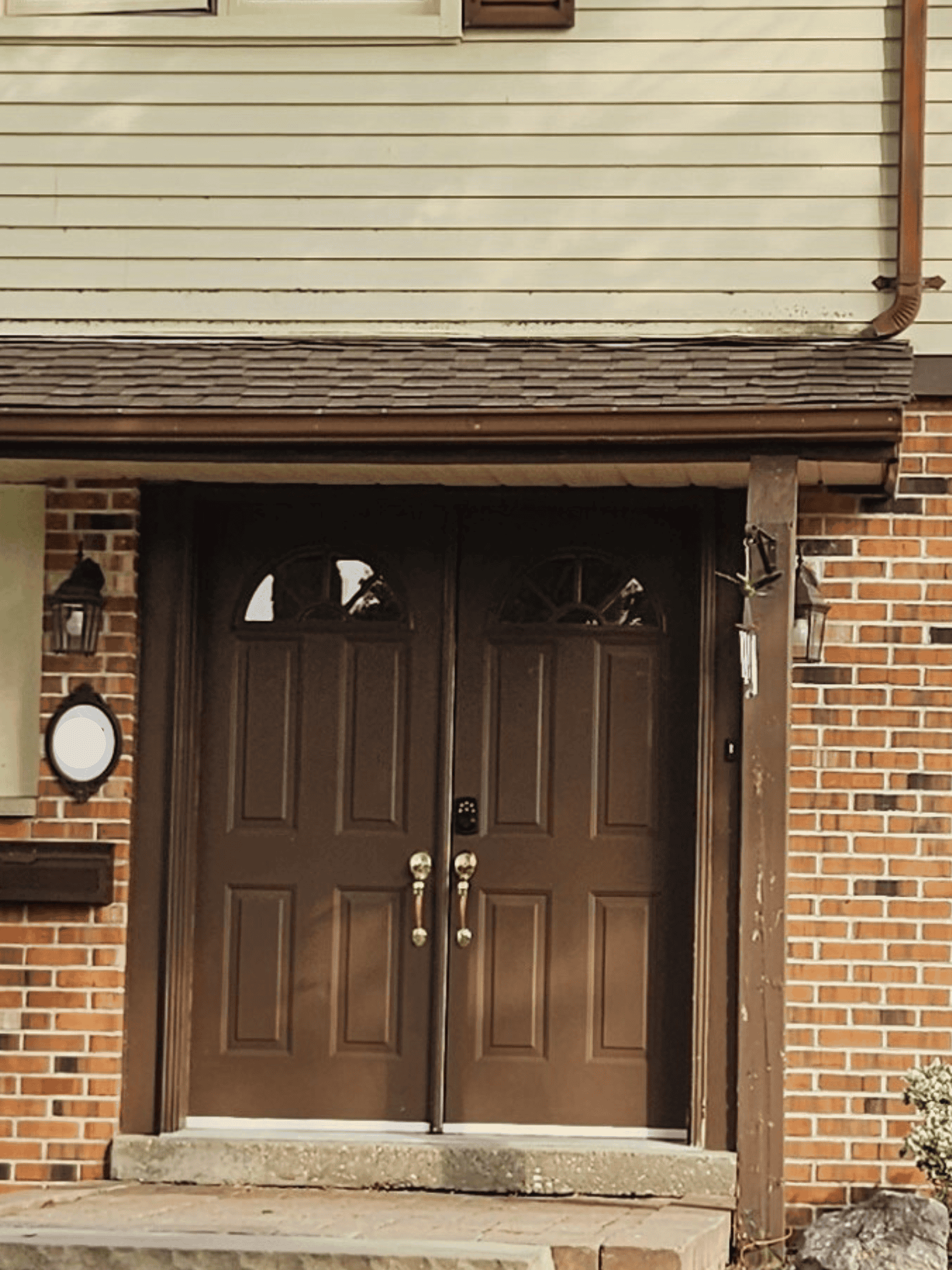 A pair of brown double front doors with gold handles, set beneath a small awning, next to a brick wall and lantern-style lights.