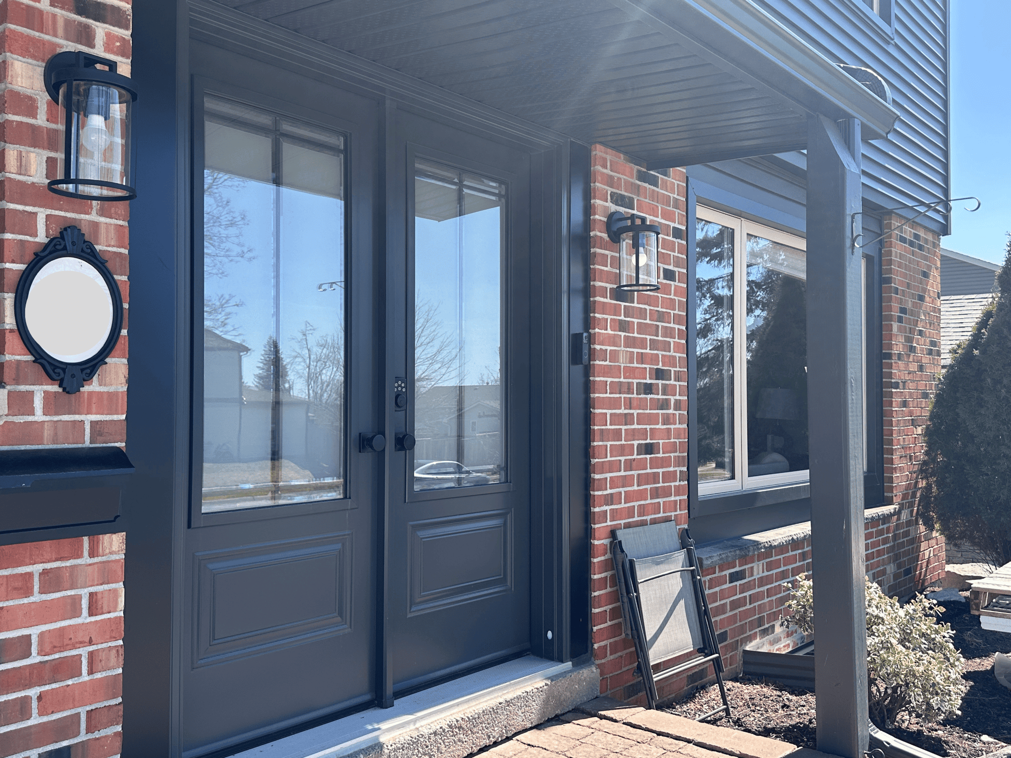 A double glass front door with dark trim on a red brick house, flanked by wall-mounted lantern lights and a small garden bed with shrubs.