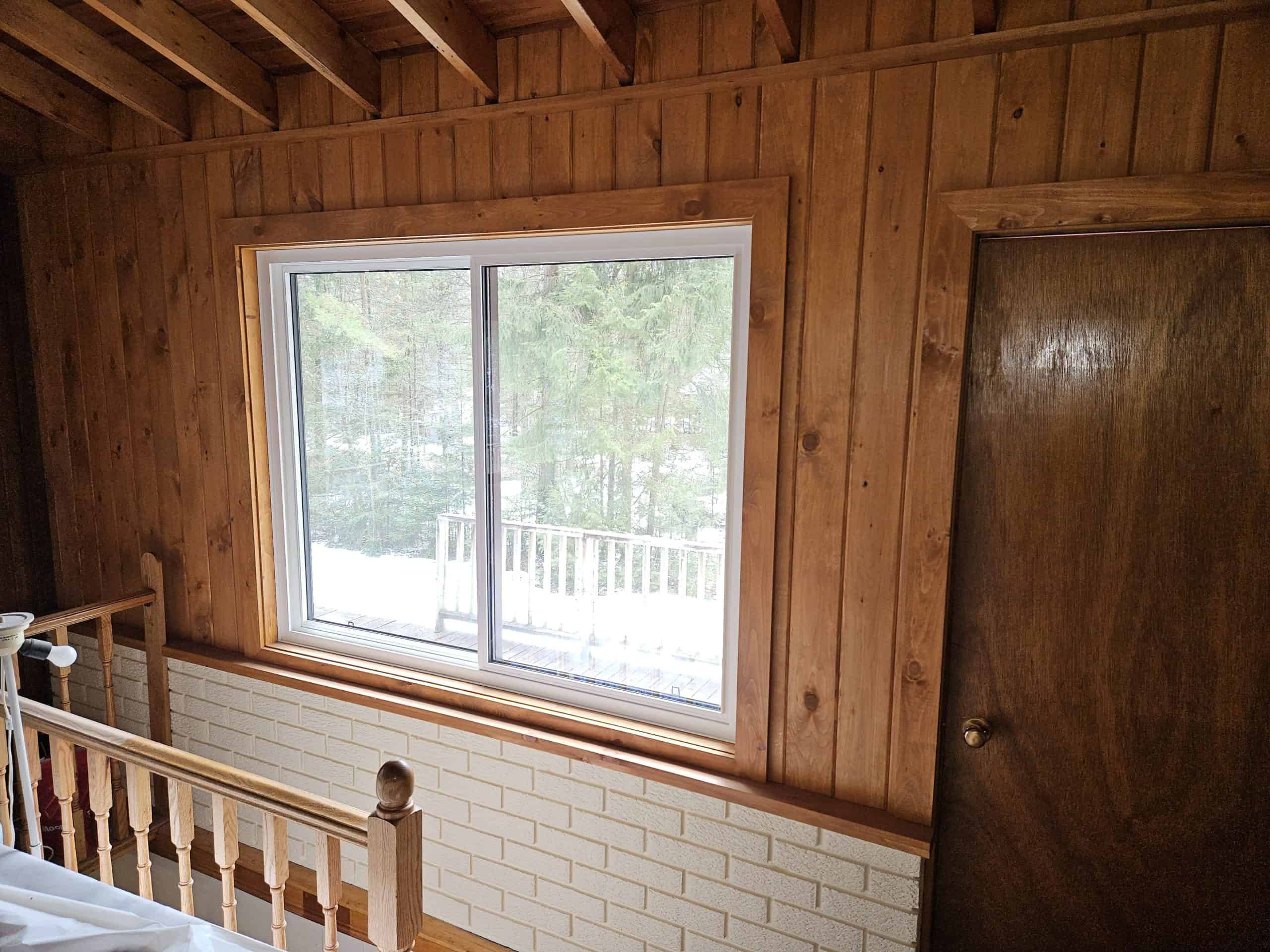 Wood-paneled room with a window overlooking trees, part of a railing visible, and a closed wooden door on the right. Home transformation