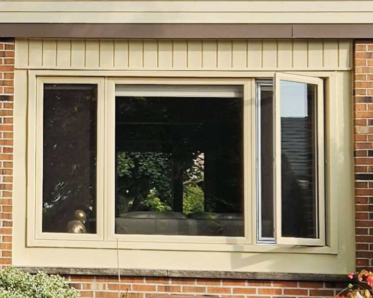 A beige-framed window set in a brick wall, with one side window panel open. Trees and part of a house roof are visible through the glass.