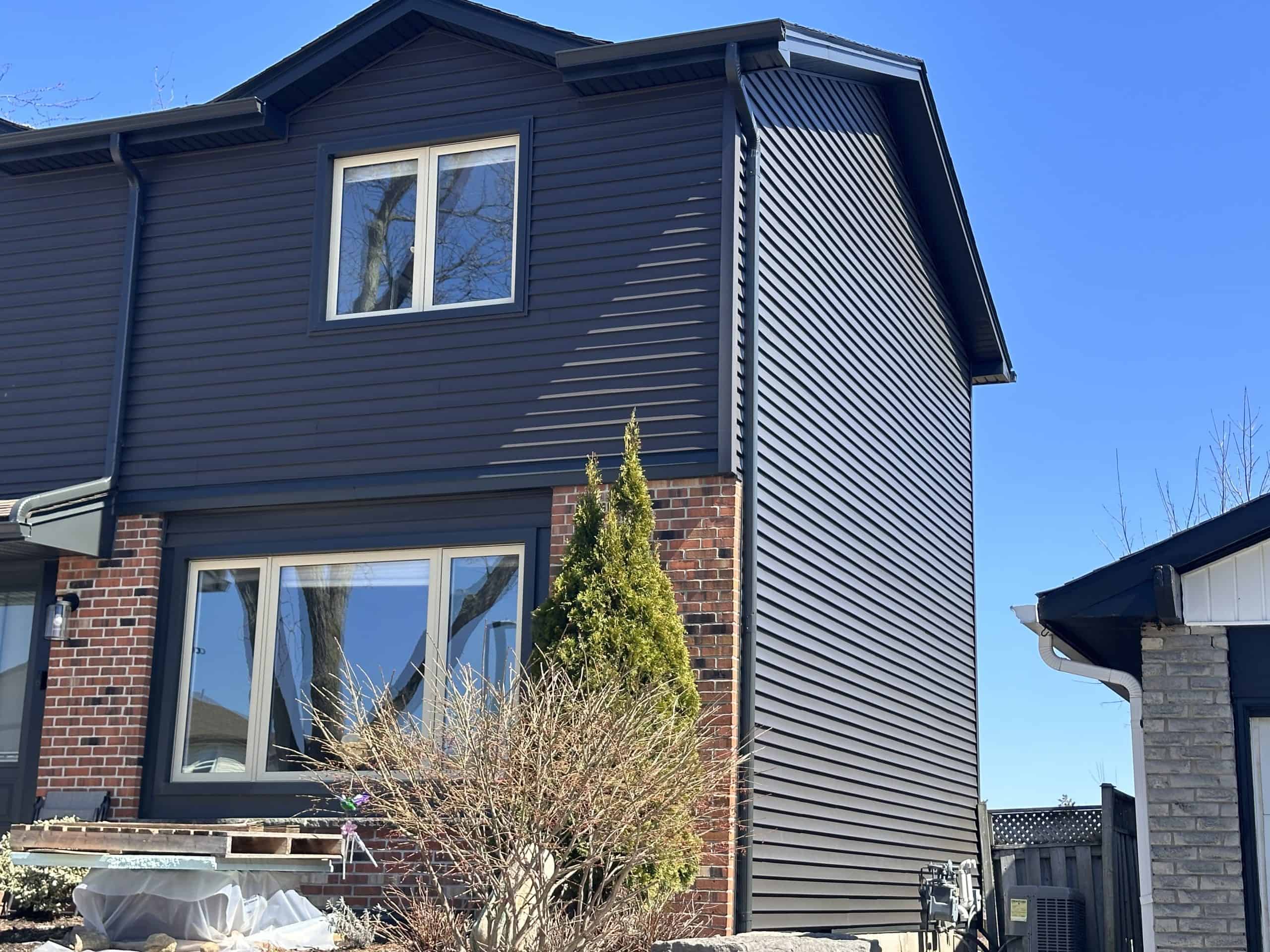 A two-story house with dark siding and red brick on the lower level, featuring large windows and a small shrub in the front yard under a clear blue sky.