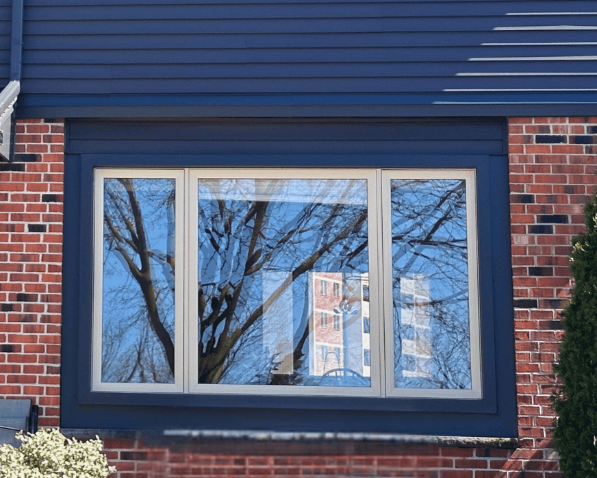 A large window on a brick house reflects leafless tree branches and sky; part of a building and objects are visible inside through the glass.